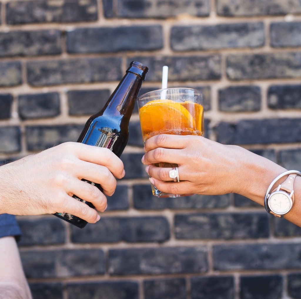 Two friends clinking a beer bottle and a glass against a brick wall